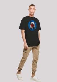Schwarzes T-Shirt mit "The Who"-Schriftzug und Logo in Rot, Weiß und Blau. Modell trägt beigefarbene Cargo-Hosen und weiße Sneakers.