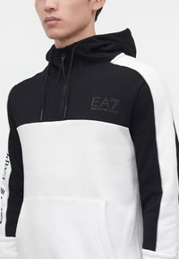 Άνδρας που φοράει μαύρο-άσπρο Emporio Armani EA7 φούτερ με φερμουάρ, τσέπη καγκουρό και λογότυπο στο στήθος και στο μανίκι.