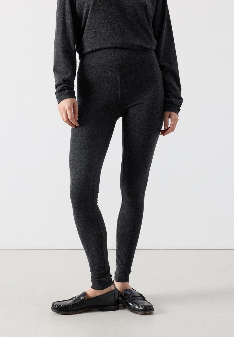 Persona que lleva leggings ajustados gris oscuro y top de manga larga a juego, combinado con mocasines negros sin cordones, de pie contra un fondo blanco.