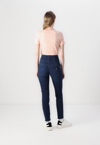 Jeansuri cu talie înaltă, de culoare albastră închisă, cu două buzunare posterior, și un detaliu mic cu logo, asortate cu un tricou roz deschis și adidași albi.