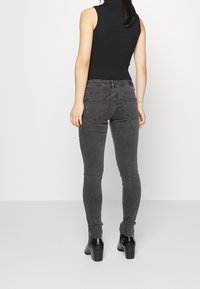 Donna che indossa jeans grigi aderenti, una maglietta nera senza maniche e stivaletti neri con tacco, appoggiata a uno sfondo bianco semplice.