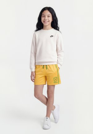 Χαμογελαστή κοπέλα με μακριά μαύρα μαλλιά φοράει λευκό φούτερ Nike, κίτρινες ποδοσφαιρικές σορτς Βραζιλίας και λευκά αθλητικά παπούτσια, στέκεται με το ένα πόδι σταυρωμένο.