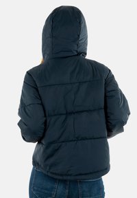 Persona che indossa una giacca a cappuccio imbottita nera e jeans blu, vista da dietro su uno sfondo bianco semplice.