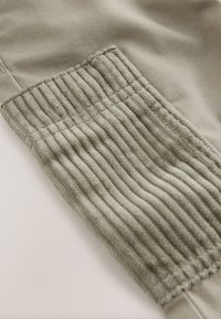 Tessuto grigio chiaro in misto cotone con pannello a coste strutturato alla caviglia. Superficie liscia con cuciture visibili e dettagli elastici.