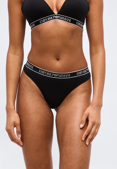 Γυναίκα που φοράει μαύρο σουτιέν και εσώρουχο σετ με το λογότυπο Emporio Armani, εμφανιζόμενη από τη μέση έως τους μηρούς πάνω σε μονόχρωμο φόντο.