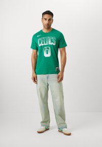Green cotton t-shirt with "CELTICS" and number "0" in white print, paired with light denim jeans and white sneakers with green accents.