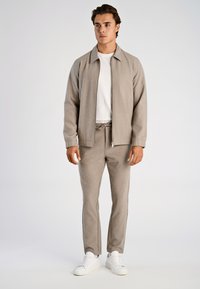 Grijze gebreide jas met een ritssluiting aan de voorkant, bijpassende grijze broek en een wit T-shirt. Heeft een ontspannen pasvorm en geribbelde manchetten. Witte sneakers.