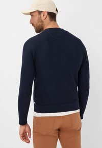 Marineblauer Strickpullover mit Rundhalsausschnitt, langen Ärmeln und geripptem Saum. Über einem weißen Hemd getragen, kombiniert mit braunen Hosen. Mit kleinem Logodetail.