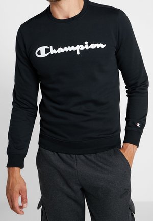 Muž v černé mikině s dlouhým rukávem s bílým logem "Champion" a tmavě šedých kalhotách, s jednou rukou v kapse, na jednobarevném pozadí.