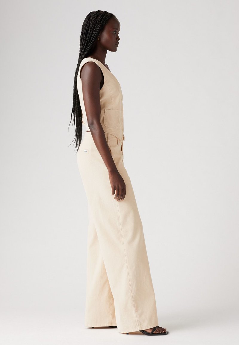 Beige Jumpsuit aus strukturiertem Stoff, mit angepasstem Oberteil, weitem Bein und zwei vorderen Taschen. Das Model steht seitlich vor einem hellen Hintergrund.