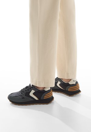 Sneaker neri e grigi con accenti bianchi, caratterizzati da una suola stratificata con strisce marroni. Le scarpe hanno una tomaia testurizzata e una punta arrotondata.