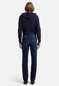 Felpa blu navy con cappuccio regolabile, abbinata a jeans di denim blu scuro con due tasche posteriori. Il modello è in piedi con le spalle rivolte verso l'osservatore.