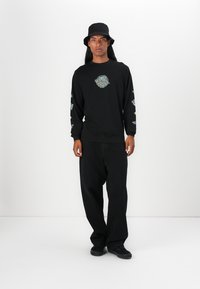 Zwart shirt met lange mouwen met een rond ontwerp op de borst en meerkleurige ontwerpen op de mouwen, gecombineerd met losse zwarte broek en een bucket hat.