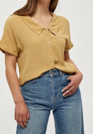Femme portant un chemisier jaune moutarde à manches courtes avec des boutons, rentré dans un jean bleu taille haute, sur un fond uni.