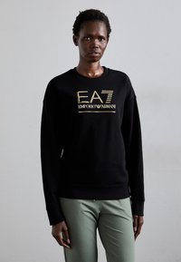 Zwarte sweatshirt met gouden logo "EA7 Emporio Armani" op de borst, met lange mouwen en geribbelde manchetten; gecombineerd met lichtgroene broek.