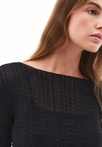 Sort tekstureret kjole med lige halsudskæring og lange ærmer, med et krøllet mønster og gennemsigtige detaljer.