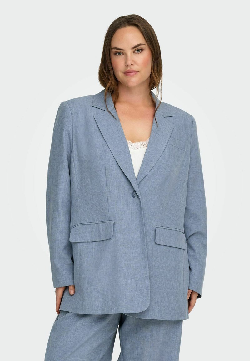 Γυναίκα που φοράει ανοιχτό μπλε υπερμεγέθη σακάκι και ταιριαστό παντελόνι πάνω από λευκή μπλούζα με δαντελωτή διακόσμηση, στέκεται μπροστά από απλό φόντο.