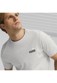 Camiseta de manga corta gris claro, fabricada con una mezcla de algodón, que presenta un logotipo negro de Puma en texto en negrita y un pequeño gráfico blanco de un puma en el pecho.