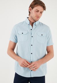 Camisa de manga corta de color azul claro con dos bolsillos en el pecho, botones negros y cuello abotonado. La tela tiene una apariencia texturizada.