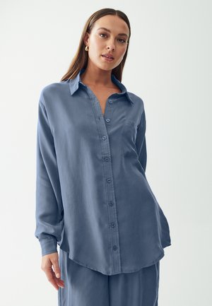 Lichtblauwe blouse met lange mouwen, een zachte textuur, knoopsluiting aan de voorzijde, kraag en een enkele borstzak. Het gladde materiaal valt iets soepel.