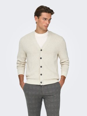 Cardigan en maille beige avec col en V, cinq boutons noirs, poignets côtelés et texture lisse, porté sur une chemise blanche et un pantalon gris.