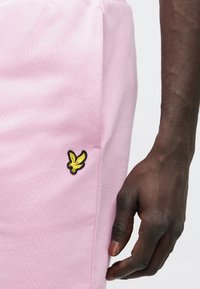 A rózsaszín melegítőnadrág texturált anyagból készült, oldalán zsebbel és a bal csípőnél sárga hímzett madár logóval. Kéz látható.