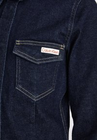 Tmavě modrá džínová bunda s knoflíkovým zapínáním, výraznou náprsní kapsou a červenou a bílou štítkovou nálepkou Calvin Klein.