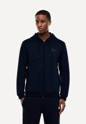 Giovane uomo che indossa una felpa nera con zip e coulisse e pantaloni neri abbinati, in piedi con le mani nelle tasche, guarda leggermente a sinistra.