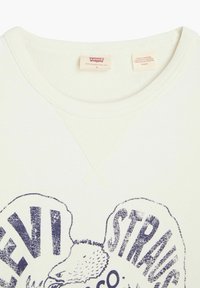 Felpa bianca sporca con grafica vintage di Levi's, colletto rotondo e texture a coste. La stampa è blu scuro con effetto invecchiato.