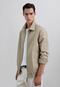Chaqueta ligera beige con cuello alto, que cuenta con un cierre de cremallera y puños elásticos. Lleva puesta una camiseta blanca. Textura suave.