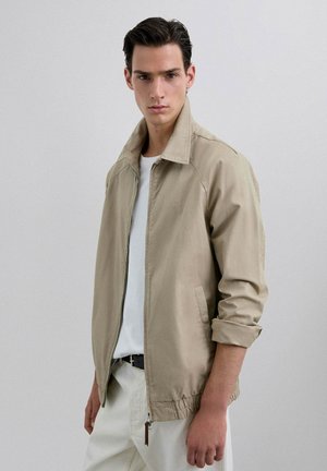 Chaqueta ligera beige con cuello alto, que cuenta con un cierre de cremallera y puños elásticos. Lleva puesta una camiseta blanca. Textura suave.