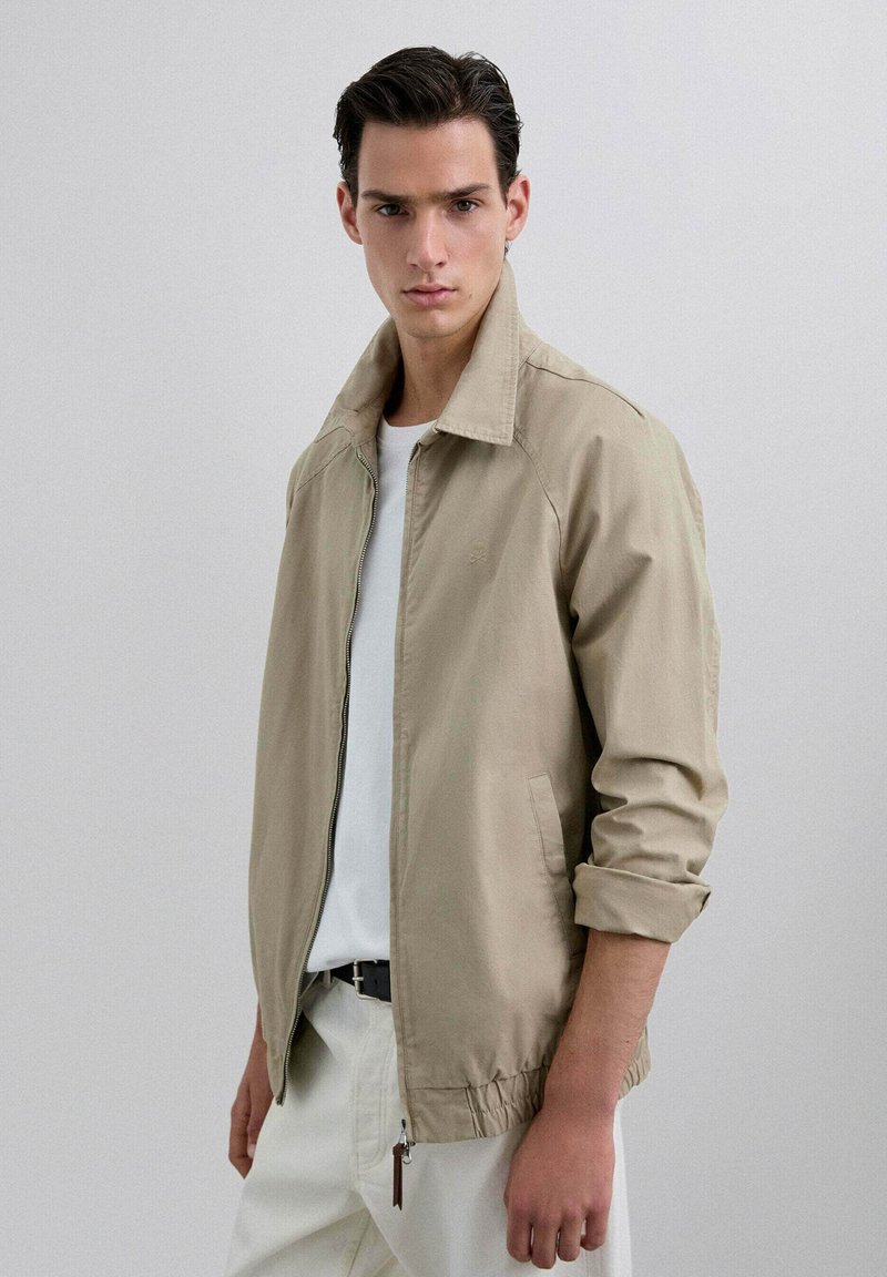 Chaqueta ligera beige con cuello alto, que cuenta con un cierre de cremallera y puños elásticos. Lleva puesta una camiseta blanca. Textura suave.