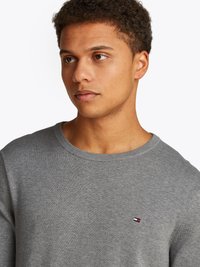Sweater cinza de gola redonda feito de tecido de malha. Apresenta um pequeno logótipo em vermelho, branco e azul no peito. Textura suave, design ajustado.