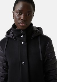 Sort frakke med et pufferjakke-design, der har blanke knapper og høj krave. Materialet kombinerer glatte og strukturerede stoffer.