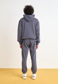 Grå hettegenser og matchende joggebukser laget av mykt stoff. Har hette, elastiske mansjetter og ankelbånd. Brukt med hvite sneakers.