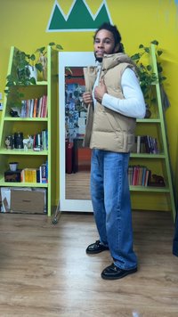 Beige gewatteerd vest met hoge kraag, gedragen over een wit shirt met lange mouwen, gecombineerd met blauwe jeans met wijde pijpen en zwarte schoenen. Groene boekenkast op de achtergrond.