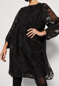 Robe noire à manches longues transparentes avec une texture florale. Silhouette fluide, encolure arrondie et taille froncée. Portée avec des collants noirs.