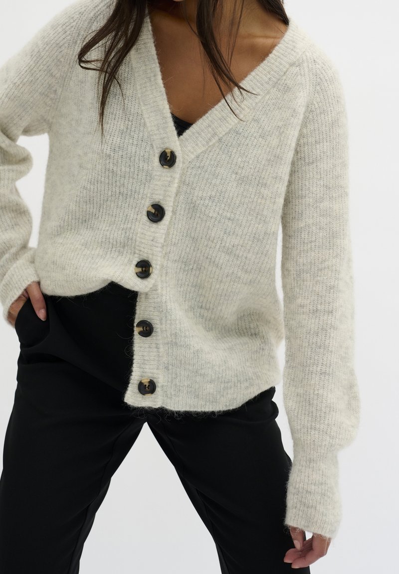 Cardigan beige clair en maille douce et texturée, avec un col en V, sept boutons noirs sur le devant et des manches amples.