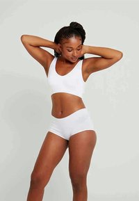 Kvinne med flette i håret som har på seg hvit sports-bh og shorts, poserer med hendene bak hodet mot en enkel lys bakgrunn.