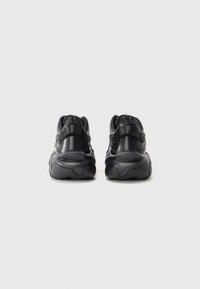 Et par svarte kraftige sneakers vist fra baksiden på en lys grå bakgrunn, som fremhever tykke såler og hælstrikker.