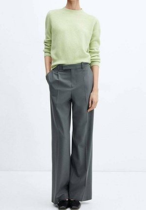 Personne portant un pull vert clair à manches longues rentré dans un pantalon à jambes larges gris taille haute avec des chaussures noires, debout devant un fond blanc.