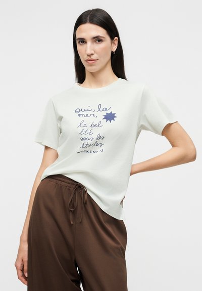 Γυναίκα με μακριά σκούρα μαλλιά, ντυμένη με λευκό μπλουζάκι με γαλλικό κείμενο και καφέ παντελόνι με κορδόνι, ποζάρει με το ένα χέρι στη μέση.