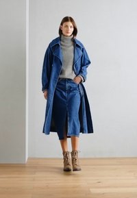 Manteau en denim sur un col roulé gris, associé à une jupe en denim avec une fente devant et des bottes à lacets marron. Sol en bois naturel.