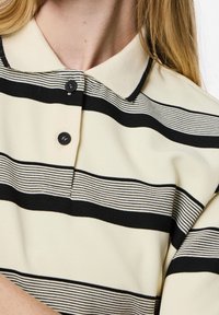 Nära bild av en krage med svart och vit randig tyg, som har tre knappar, mot en ljus bakgrund.