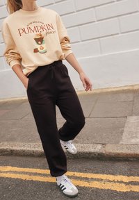 Beige sweatshirt met grafische tekst en pompoenontwerp, gecombineerd met zwarte joggers. Witte sneakers met grijze en blauwe accenten. Straatomgeving.