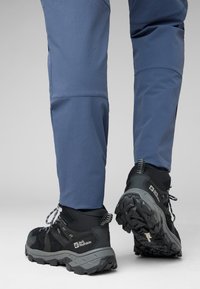 Scarpe da trekking nere con dettagli grigi, caratterizzate da una suola testurizzata e lacci riflettenti, abbinate a pantaloni blu, che mostrano un design robusto.
