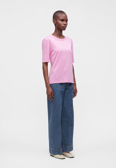 Femme debout portant un t-shirt rose à manches courtes, un jean bleu à jambes larges et des chaussures blanches à bout pointu sur un fond clair uni.