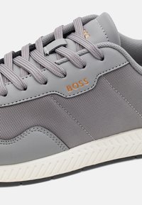Grau Sneakers mit strukturiertem Stoff und glatten Lederelementen. Ausgestattet mit grauen Schnürsenkeln, "BOSS"-Logo und einer weißen Gummisohle mit Profil.