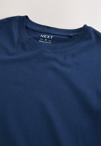 Tmavomodré tričko s okrúhlym výstrihom z mäkkej bavlny. Na prednej strane je štítok s nápisom "NEXT" a veľkosťou "M" pre bežný strih. Hladká textúra.