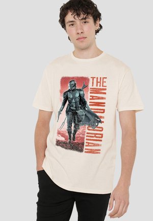 Young man wearing beige t-shirt with graphic of armored character walking and "The Mandalorian" text in red on front.